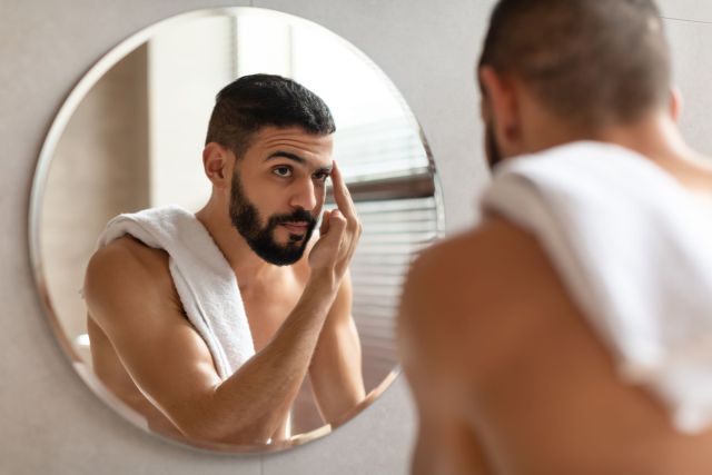 Confident young guy looking in the mirror, touching eyebrow